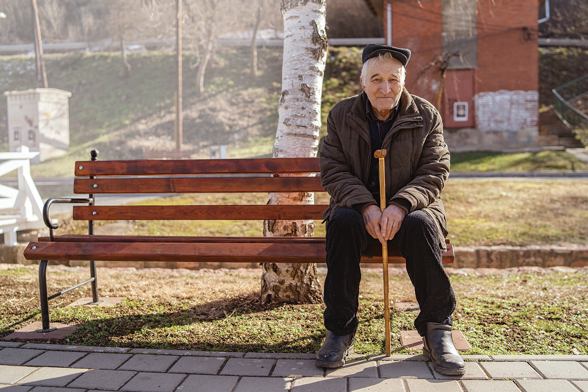 Senior old man caucasian pensioner grandfather holding his cane on the sitting on the bench by sidewalk pavement in park sunny winter or spring day looking to the camera wearing coat jacket and hat